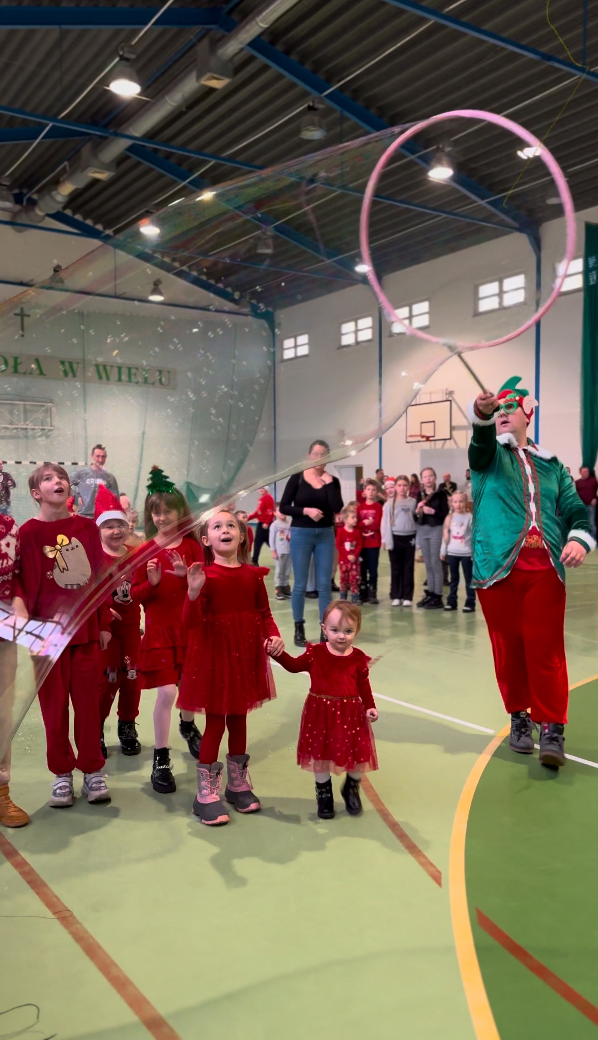 Animator w stroju elfa puszcza ogromne bańki mydlane dla grupy dzieci ubranych na czerwono w sali gimnastycznej, widoczne kosze do koszykówki i okna.