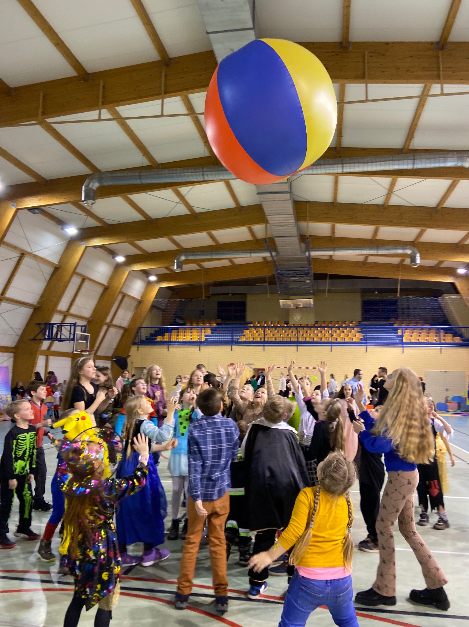 Duża grupa dzieci w kostiumach bawi się w hali sportowej, podrzucając do góry dużą, kolorową piłkę.