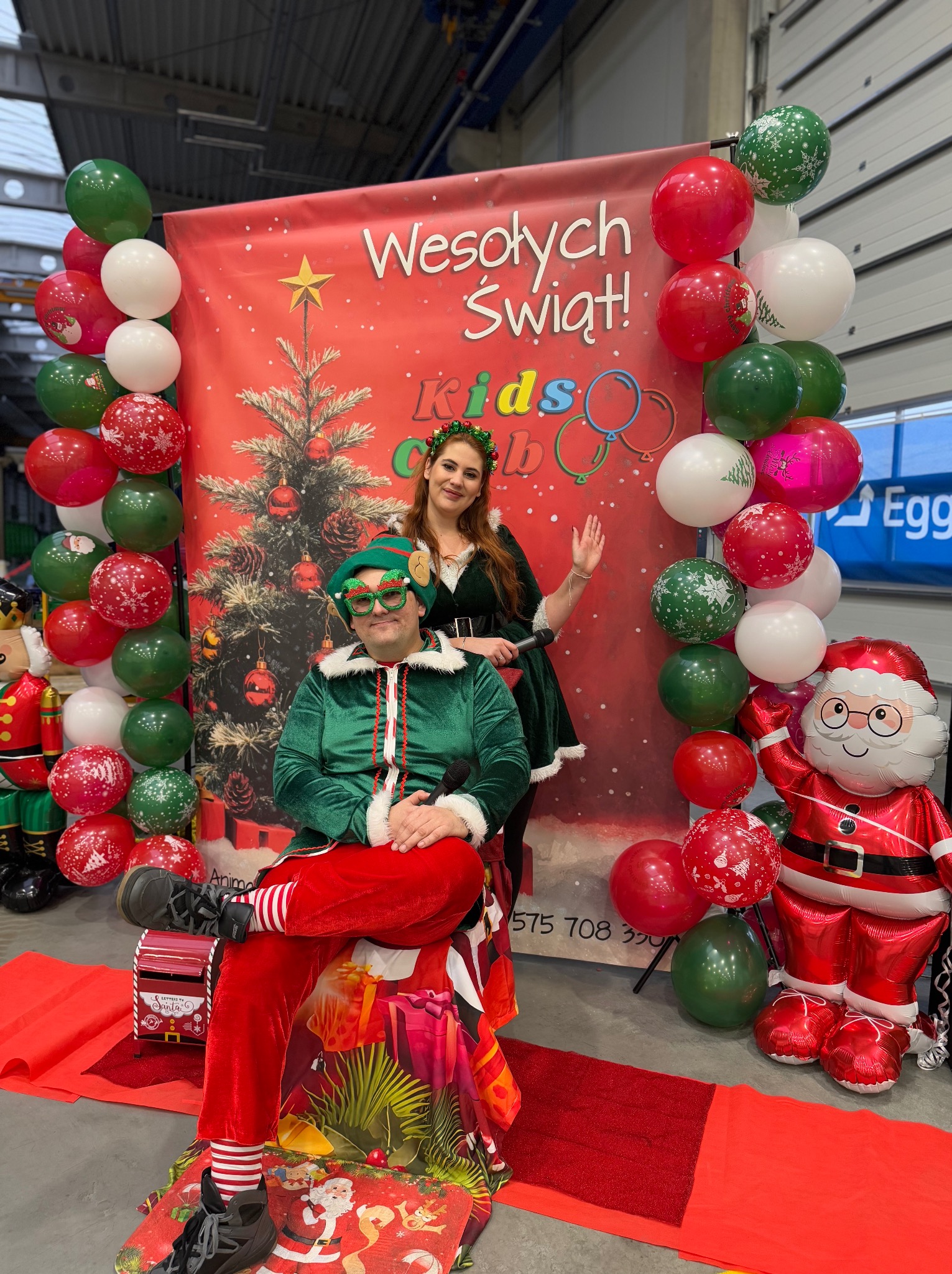 Animator w przebraniu elfa siedzi na ozdobnym krześle, obok stoi kobieta w stroju pomocnika Świętego Mikołaja, w tle dekoracje świąteczne i balony.