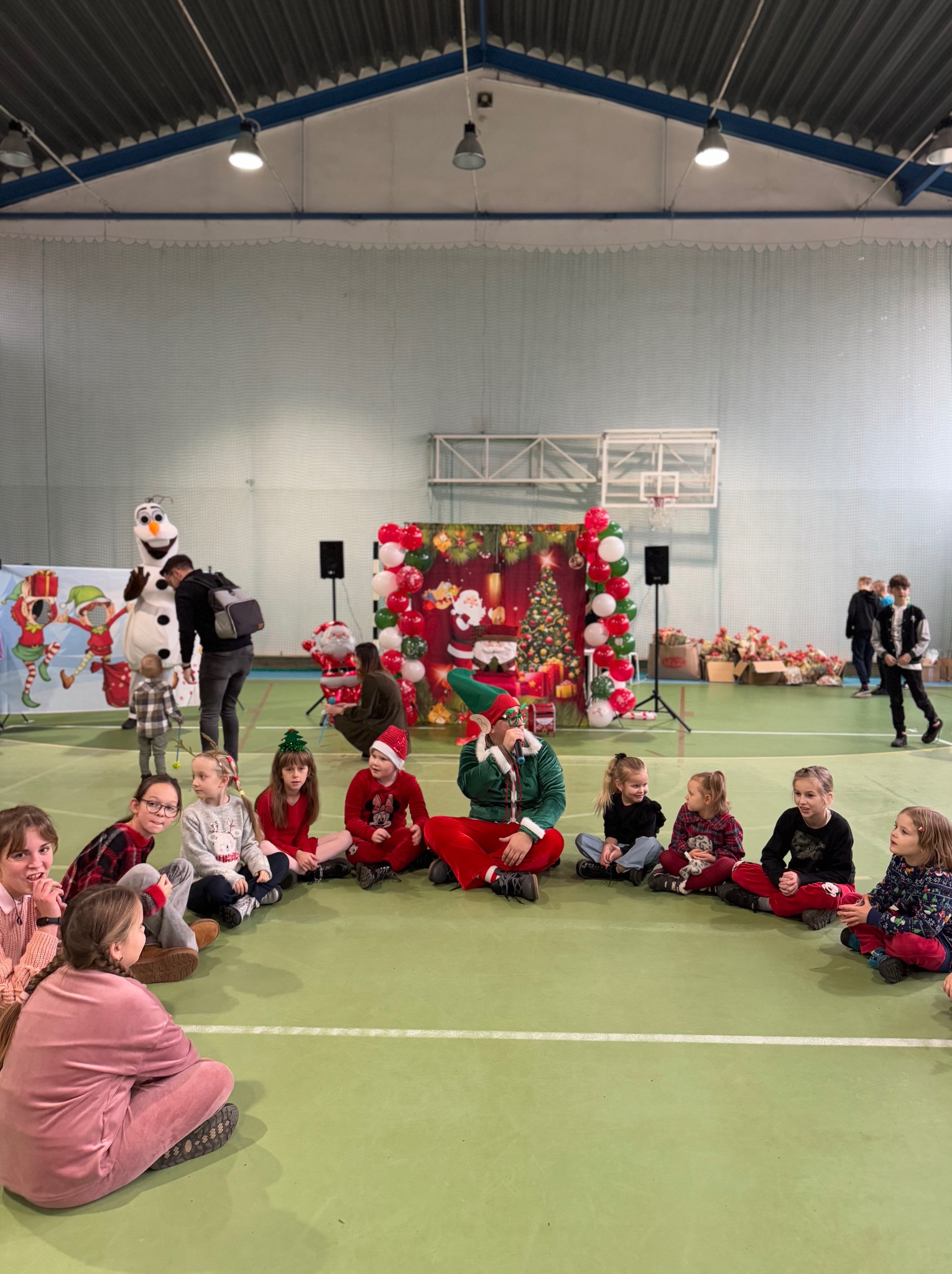 Grupa dzieci siedzi na podłodze w sali gimnastycznej, otoczona dekoracjami świątecznymi i postacią elfa z mikrofonem, w tle widoczny Olaf, balonowa dekoracja i scena z motywem świątecznym.