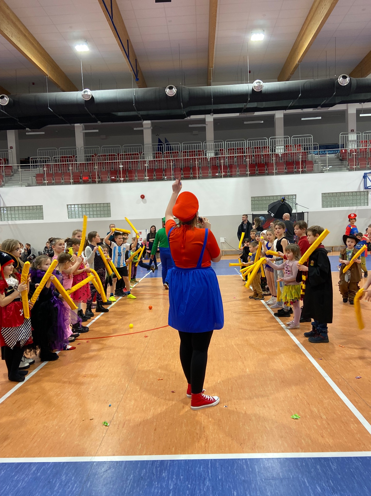 Animator w stroju postaci z gier wideo, prowadzi zabawę z dziećmi przebranymi w kostiumy, trzymającymi żółte rekwizyty, w hali sportowej.