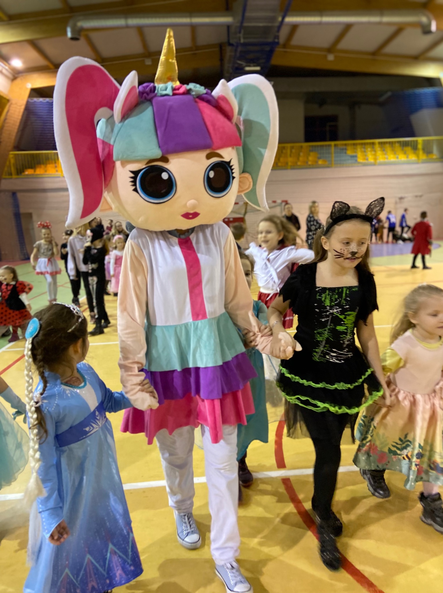 Animator w kostiumie jednorożca z dużymi oczami prowadzi grupę dzieci przebranych w kostiumy na tle sali gimnastycznej.