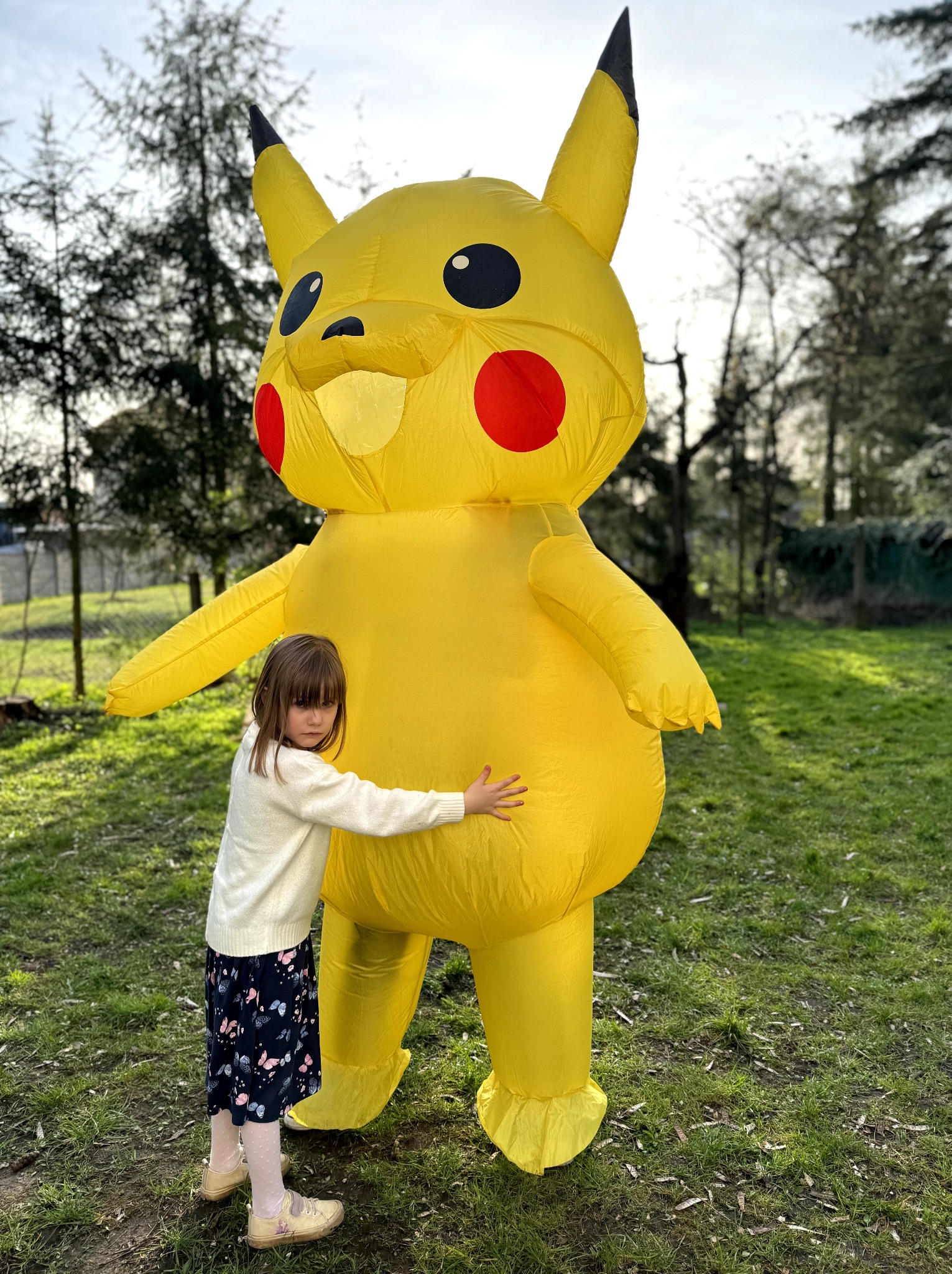 Dziewczynka w jasnym swetrze i wzorzystej spódnicy przytula dmuchany kostium Pikachu na trawniku w plenerze.