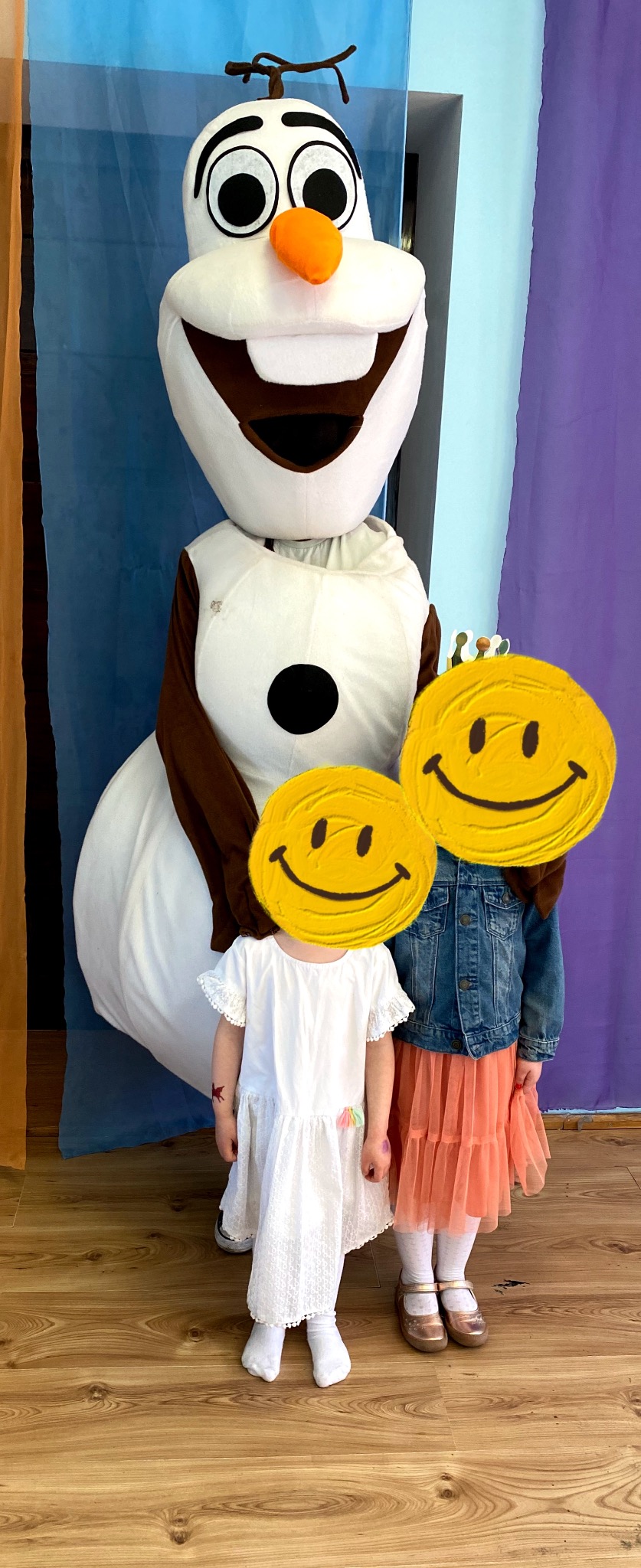 Dwie dziewczynki pozują z animatorem przebranym za Olafa z Krainy Lodu na tle dekoracji w odcieniach niebieskiego i fioletowego.