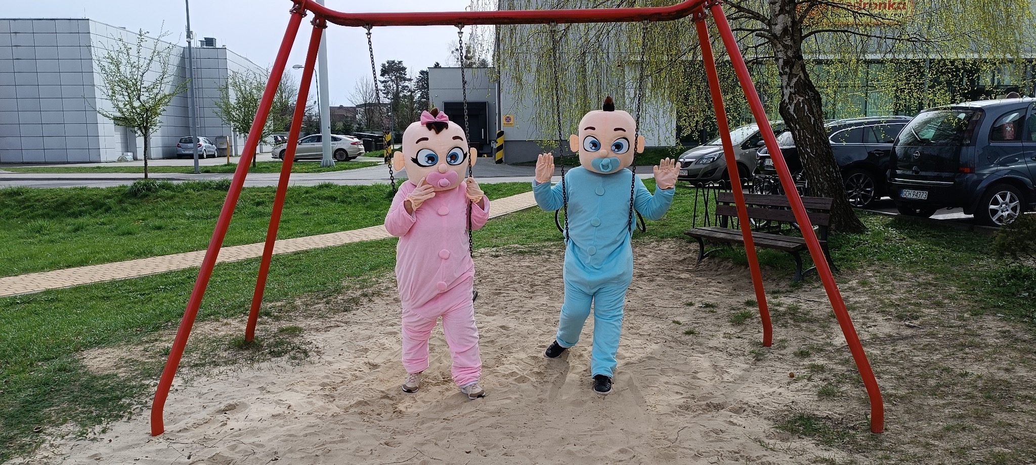 Dwie osoby w kostiumach niemowląt, różowym i niebieskim, stojące w piaskownicy pod czerwonym huśtawką na tle drzew i parkingu.