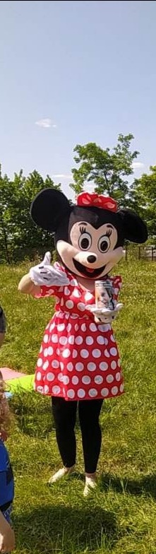 Animator w stroju Minnie Mouse w czerwonej sukience w białe grochy, stojący na trawie z napojem, wykonujący gest kciuka w górę.