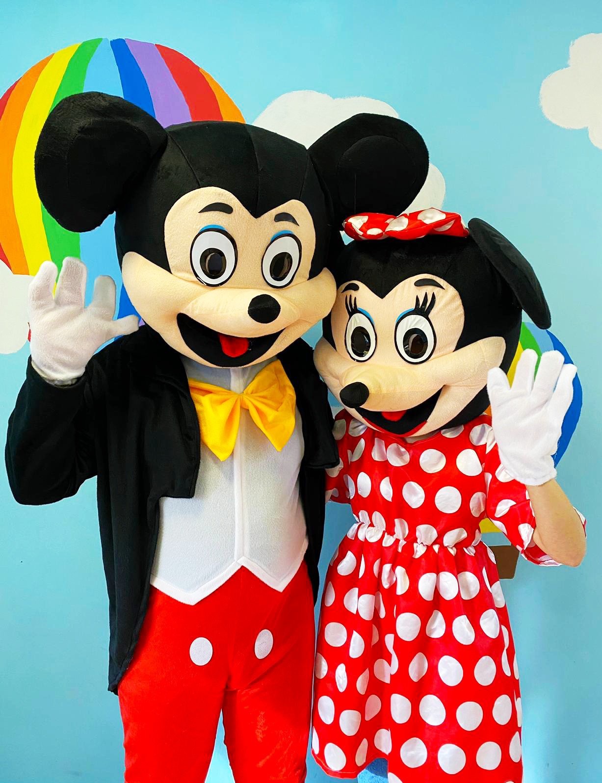 Dwie osoby w kostiumach Myszki Miki i Minnie machają rękami na tle błękitnej ściany z tęczą i chmurami.