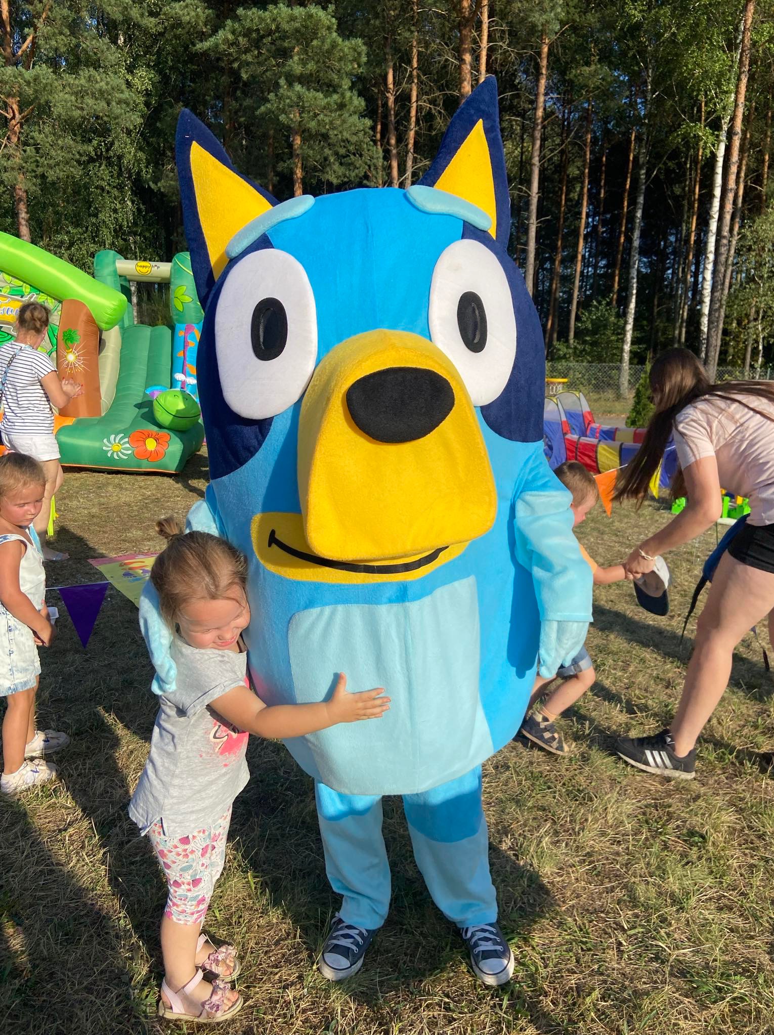 Mała dziewczynka w różowych sandałach przytula się do dużego, niebieskiego pluszowego kostiumu postaci z bajki Bluey na tle dmuchanego zamku i drzew podczas imprezy plenerowej.
