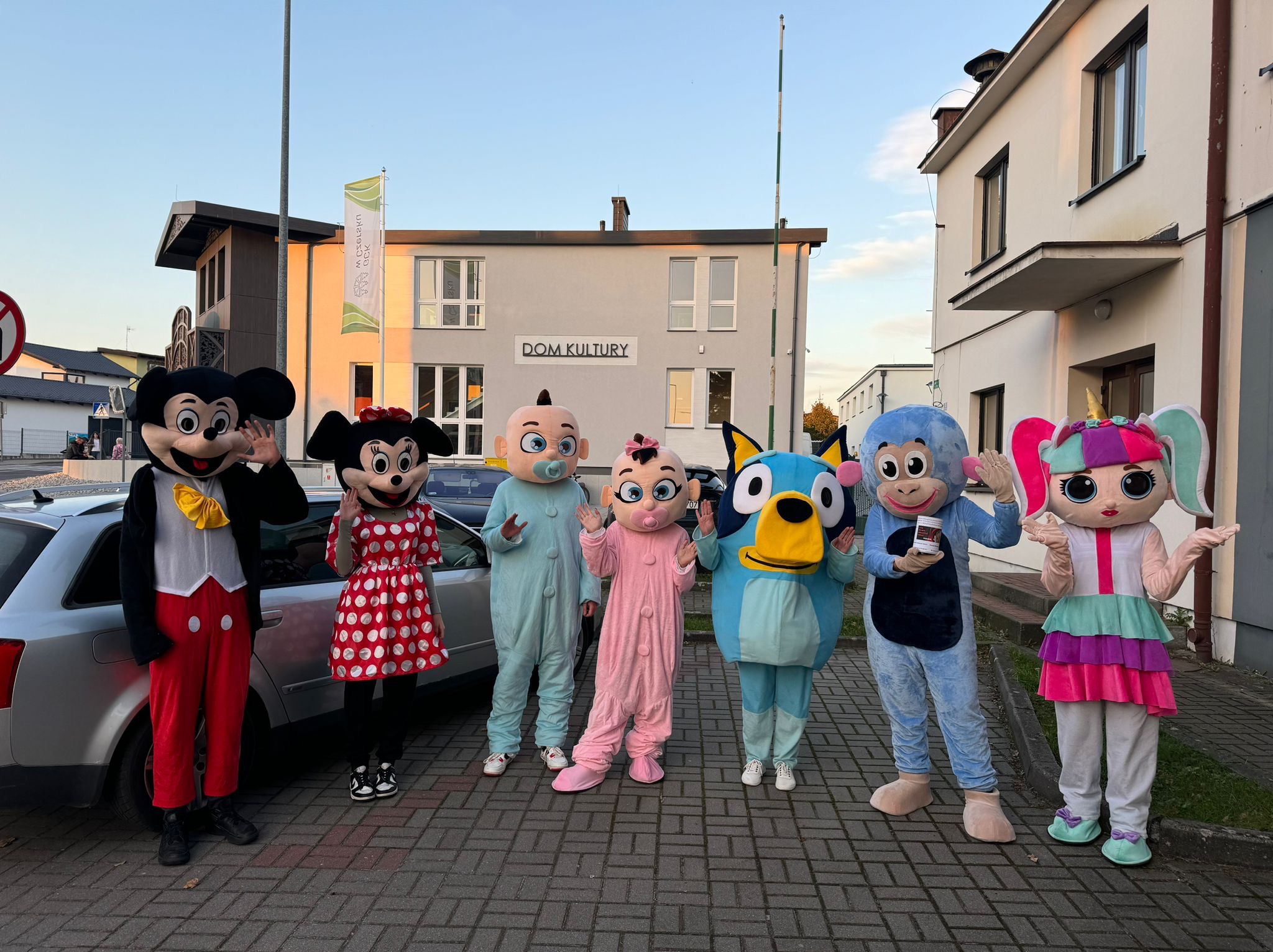 Grupa osób w kostiumach postaci z bajek, w tym Myszka Miki i Minnie, ustawiona przed budynkiem Domu Kultury i zaparkowanym samochodem, prezentująca się w pełnym słońcu.