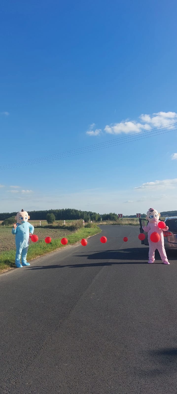 Dwie osoby w kostiumach niemowląt, niebieskim i różowym, stoją na asfalcie z czerwonymi balonami, na tle zielonego krajobrazu i błękitnego nieba.