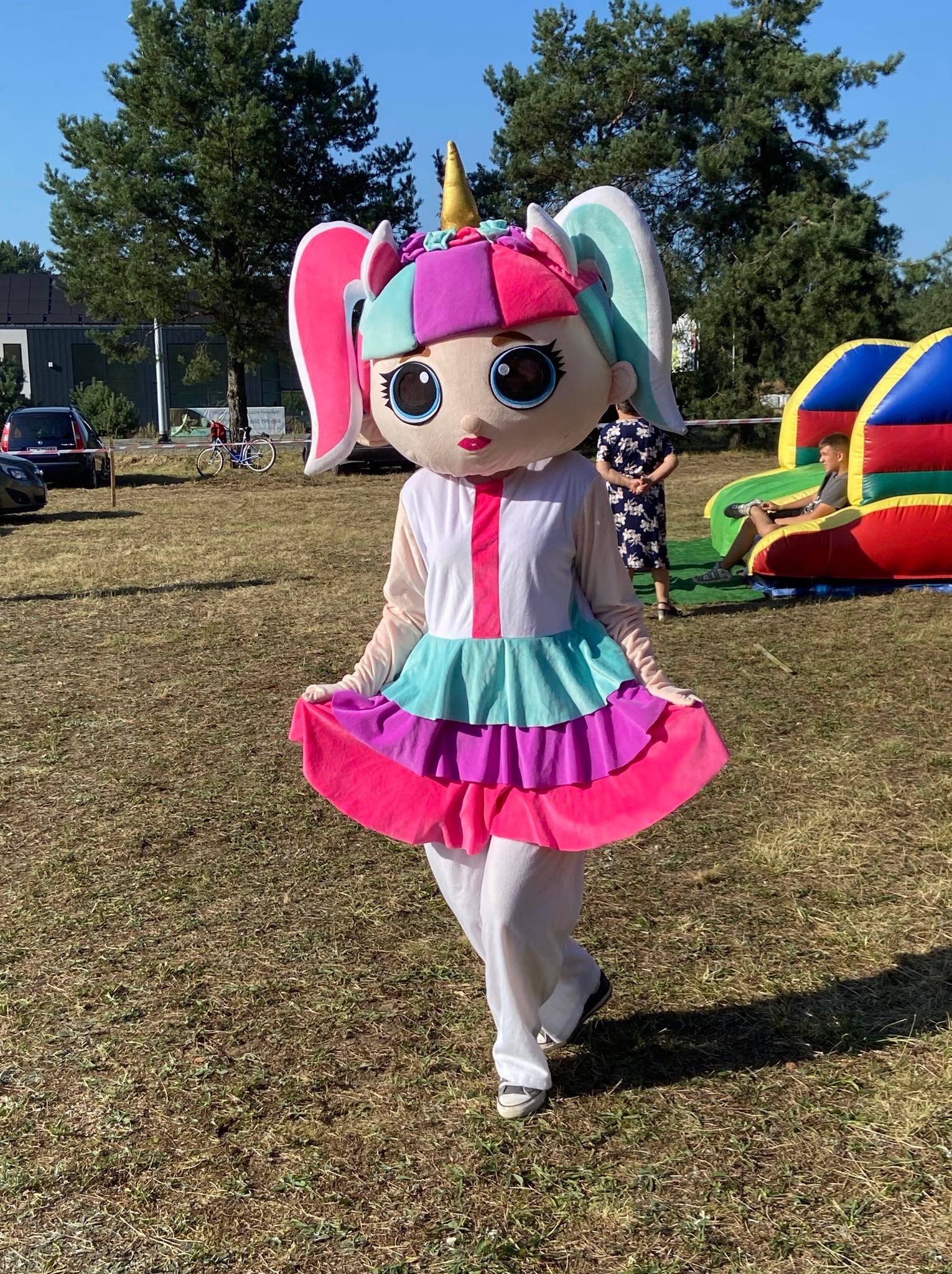 Osoba w kostiumie jednorożca z dużymi oczami i kolorową sukienką, stojąca na trawie, w tle dmuchany zamek i ludzie.