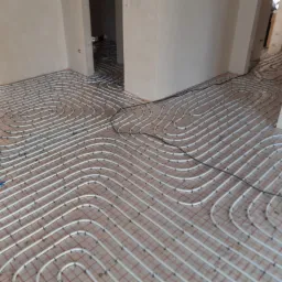 Rozłożone rury ogrzewania podłogowego na podkładzie z siatki w surowym wnętrzu, widoczne przejścia do kolejnych pomieszczeń.