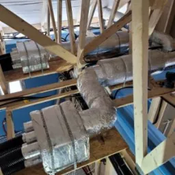 Montaż rekuperacji na poddaszu: widoczne rury wentylacyjne owinięte folią aluminiową, czarne rury doprowadzające powietrze, drewniana konstrukcja dachu w tle, elementy niebieskiej blachy trapezowej.