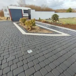 Ułożona szara kostka brukowa z białymi obramowaniami rabat i ścieżki prowadzącej do bramy. W tle częściowo widoczny biały bus Volkswagen i ogrodzenie.