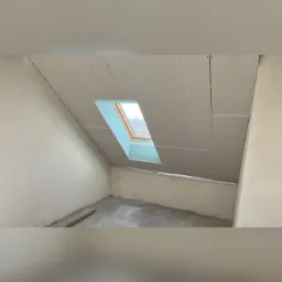 Poddasze w trakcie ocieplania: widoczne płyty kartonowo-gipsowe na skosach dachu i ścianach, okno dachowe z błękitną ramą, surowy beton na podłodze.