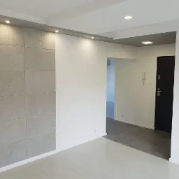 Nowoczesny przedpokój z betonową ścianą dekoracyjną z podświetleniem punktowym, szarą podłogą w przejściu i białą podłogą w dalszej części, czarne drzwi wejściowe.