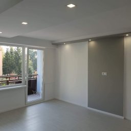 EWELINA MIKOŁAJCZYK BEST HOME - Jasne, nowocześnie wykończone wnętrze pokoju z panelami podłogowymi, świeżo pomalowanymi ścianami w odcieniach bieli i szarości, z oknem balkonowym wychodzącym na zieleń.