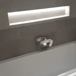 Szare płytki w łazience z dekoracyjnym paskiem mozaiki, wnęka z oświetleniem LED nad białą wanną i chromowaną baterią wannową z prysznicem.