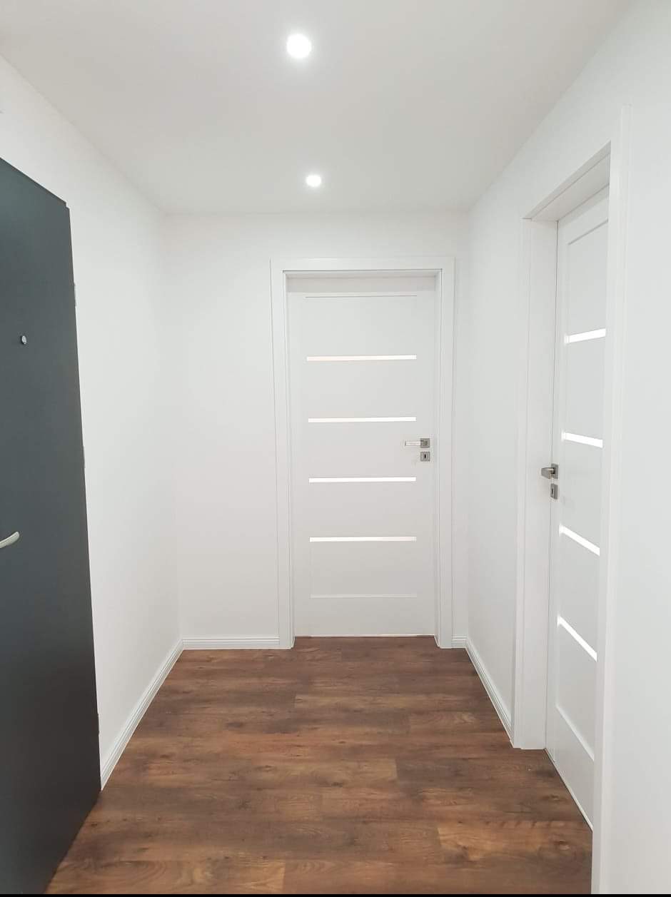 Wnętrze korytarza z brązowymi panelami podłogowymi, białymi ścianami i dwiema nowoczesnymi białymi drzwiami z poziomymi przeszkleniami, oświetlone punktowymi lampami sufitowymi.