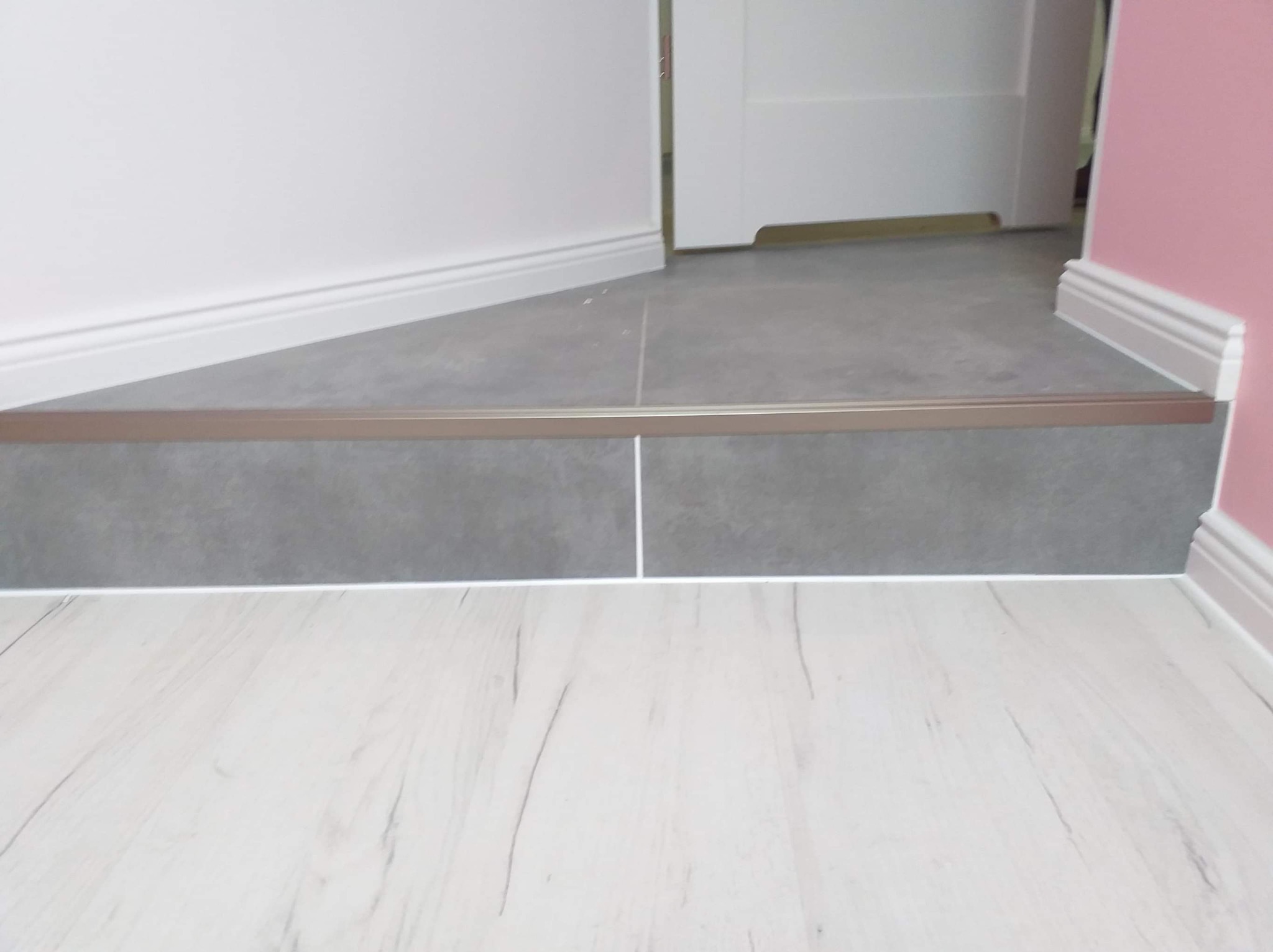 Połączenie jasnych paneli podłogowych z szarymi płytkami podłogowymi, zabezpieczone metalową listwą progową w odcieniu brązu, widoczne białe listwy przypodłogowe przy ścianie w kolorze różowym...