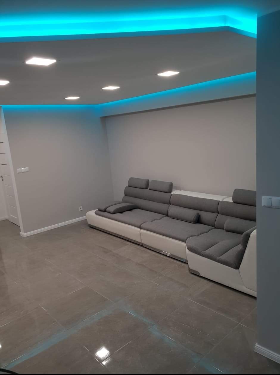 Wykończone wnętrze salonu w Ostrołęce z szarą, narożną kanapą, szarą podłogą z płytek imitujących kamień i podwieszanym sufitem z oświetleniem LED.