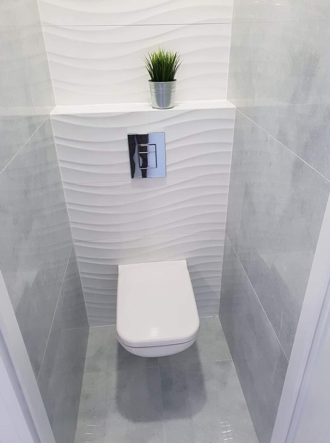 Nowoczesna toaleta podwieszana w szaro-białej łazience z dekoracyjnymi, falowanymi płytkami na ścianie i metalową doniczką z trawą na półce.