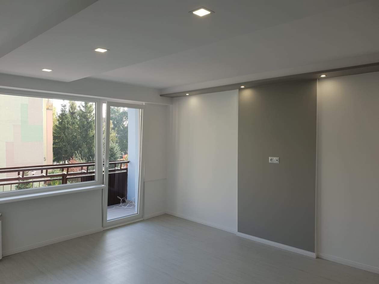 Jasne, nowocześnie wykończone wnętrze pokoju z panelami podłogowymi, świeżo pomalowanymi ścianami w odcieniach bieli i szarości, z oknem balkonowym wychodzącym na zieleń.