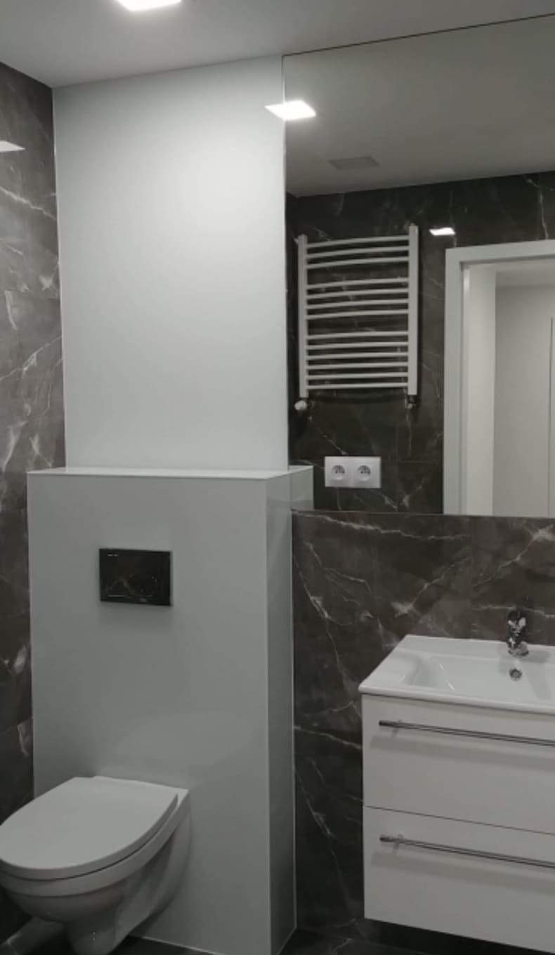 Nowoczesna łazienka w odcieniach szarości z marmurowym wzorem, wisząca toaleta z zabudową, biała umywalka z szafką i grzejnik drabinkowy.