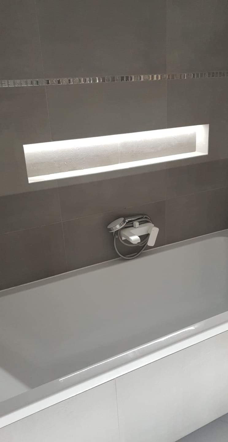 Szare płytki w łazience z dekoracyjnym paskiem mozaiki, wnęka z oświetleniem LED nad białą wanną i chromowaną baterią wannową z prysznicem.
