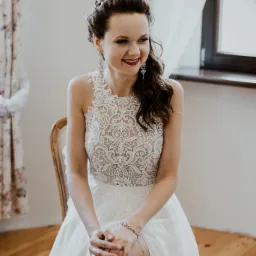 Uśmiechnięta panna młoda w koronkowej sukni siedzi na krześle, z widocznym upięciem włosów, kolczykami i delikatnym makijażem ślubnym.