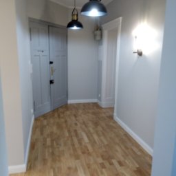 MIKO&GRESS - Wnętrze korytarza po remoncie: świeżo pomalowane ściany w odcieniach szarości, drewniana podłoga ułożona w jodełkę, wiszące lampy w stylu industrialnym i kinkiet na ścianie.