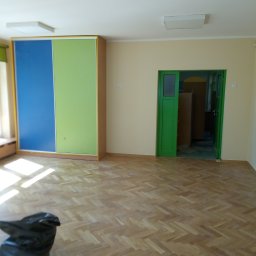 MIKO&GRESS - Pusty pokój z parkietem w jodełkę, świeżo pomalowanymi ścianami w kolorze kremowym, zielonymi drzwiami i szafą z przesuwanymi drzwiami w kolorach zielonym i niebieskim. Na podłodze leży czarny...