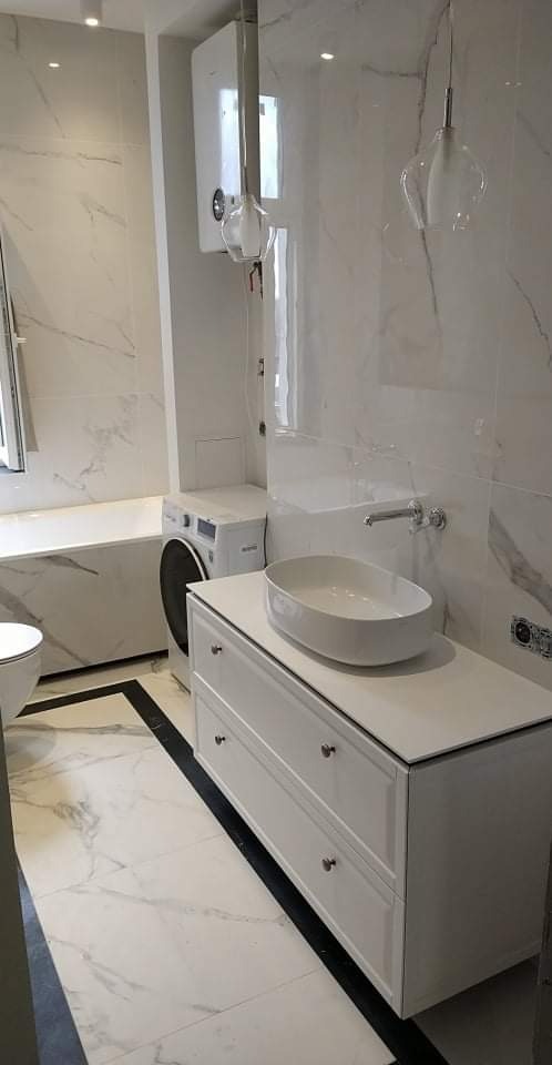 Wykończona łazienka z marmurowymi płytkami, białą umywalką nablatową na szafce z szufladami, pralką, bojlerem i wiszącymi lampami.