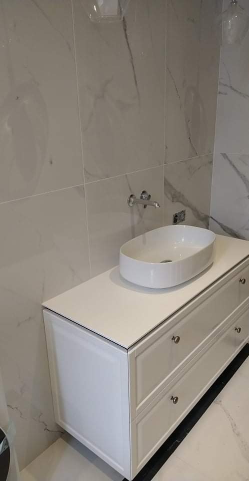 Biała umywalka nablatowa o owalnym kształcie na szafce z trzema szufladami, marmurowe płytki na ścianie i podłodze, chromowana bateria umywalkowa.