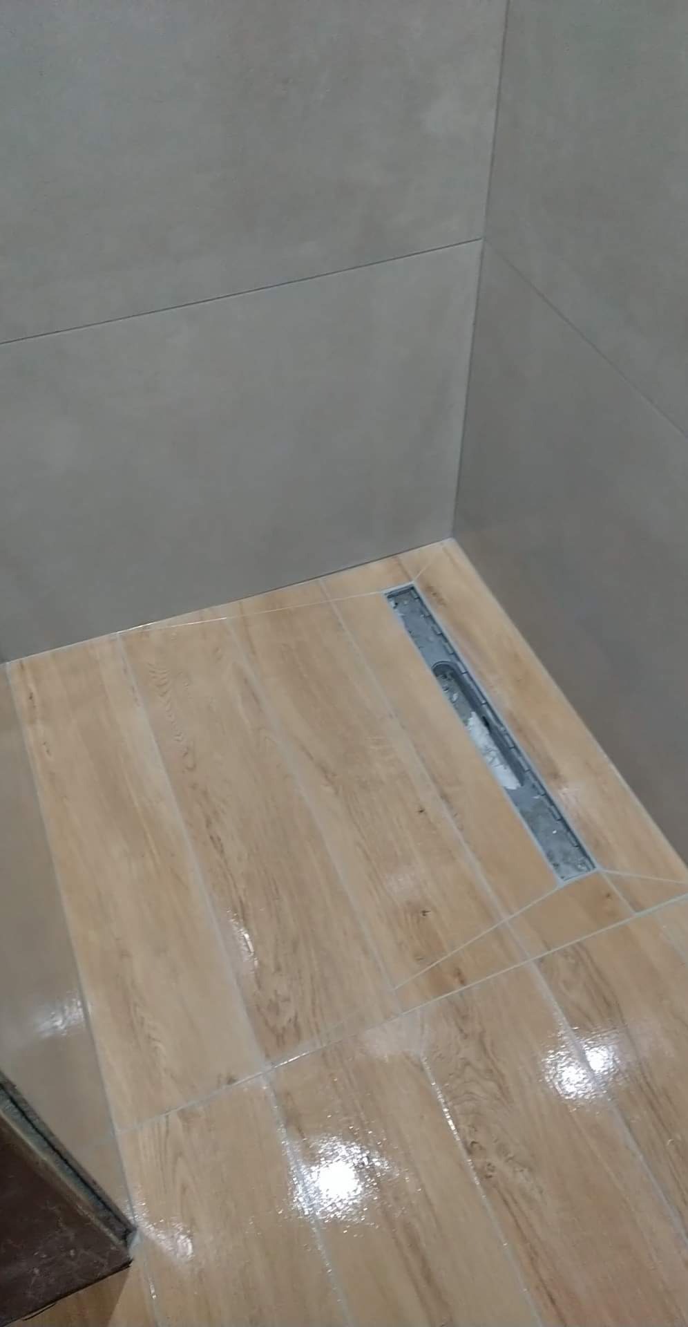 Ukończona podłoga w kabinie prysznicowej z odpływem liniowym, wyłożona jasnymi płytkami imitującymi drewno, z szarymi płytkami na ścianach.