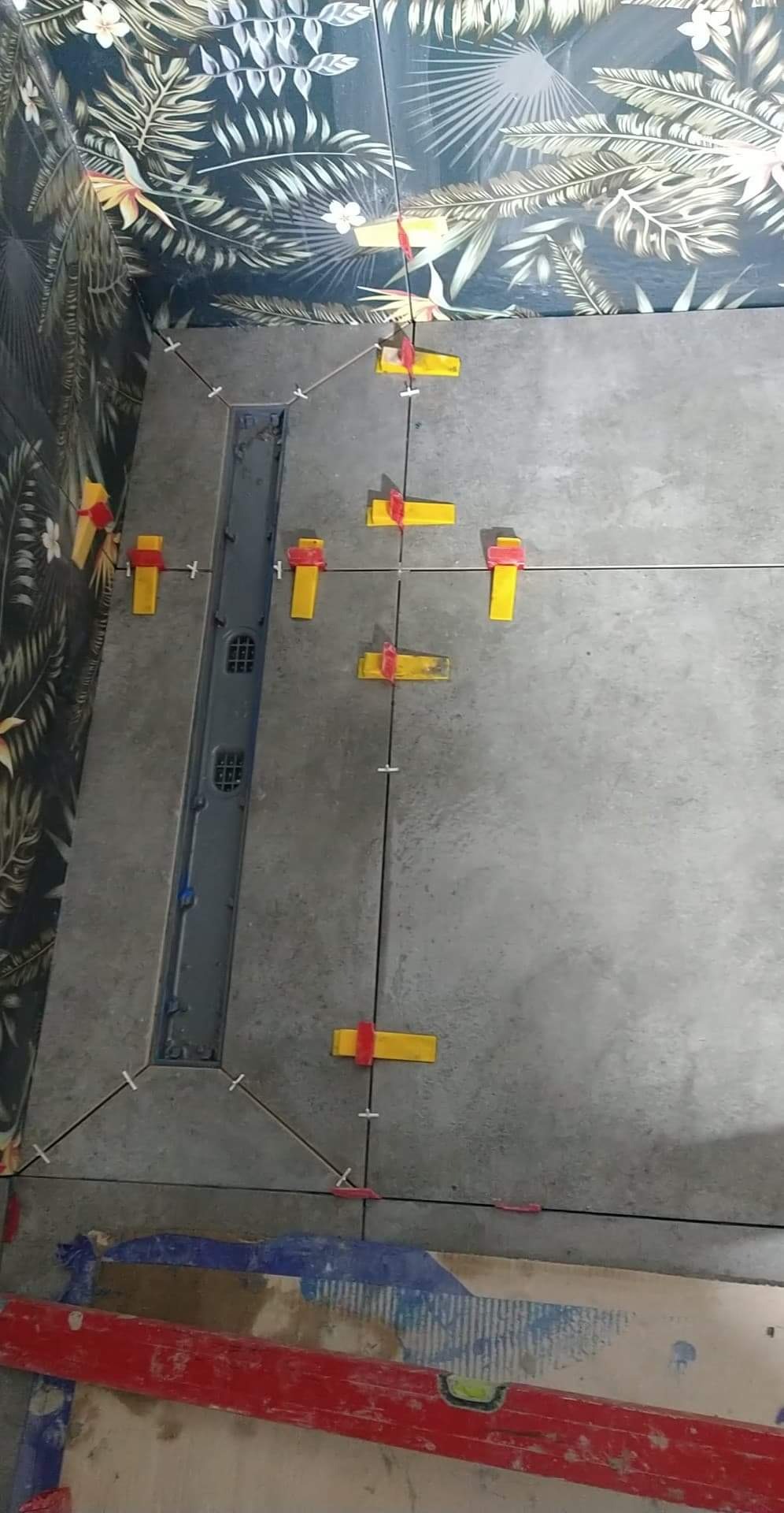 Szare płytki podłogowe w trakcie układania, z widocznym odpływem liniowym i systemem poziomowania płytek, na ścianie wzorzyste płytki imitujące liście.