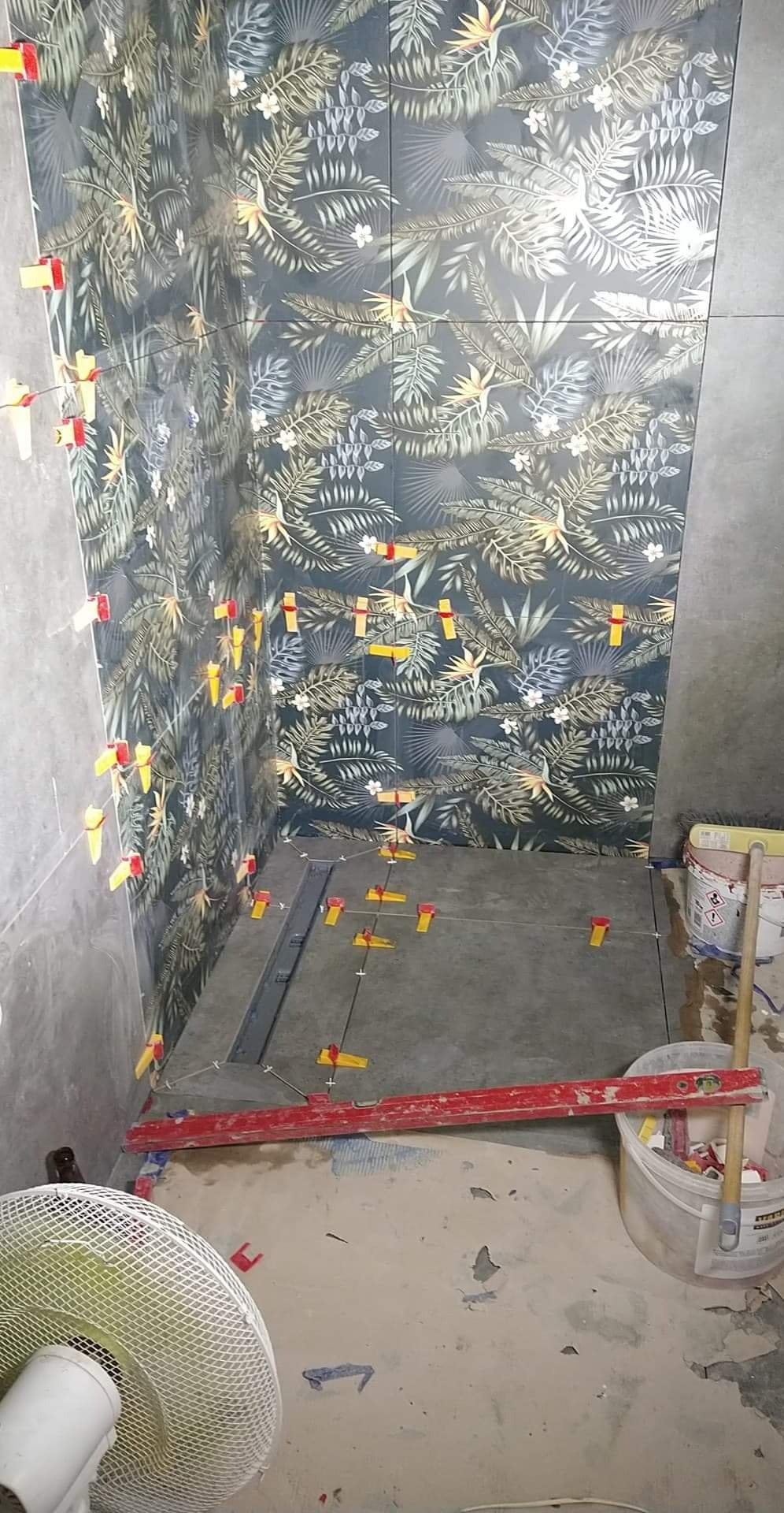 Łazienka w trakcie układania płytek z motywem tropikalnym na ścianach, widoczne krzyżyki dystansowe, poziomica, wiadro z zaprawą i wentylator.