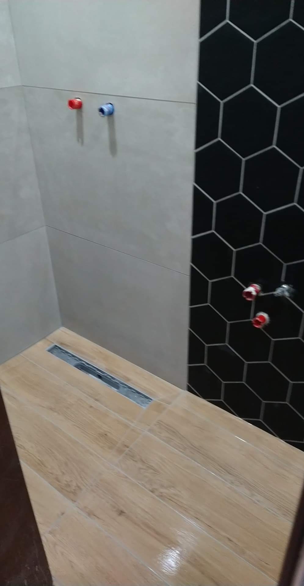 Nowoczesna kabina prysznicowa z drewnopodobnymi płytkami na podłodze, odpływem liniowym i ścianą z szarych płytek oraz fragmentem ściany z czarnych heksagonów.