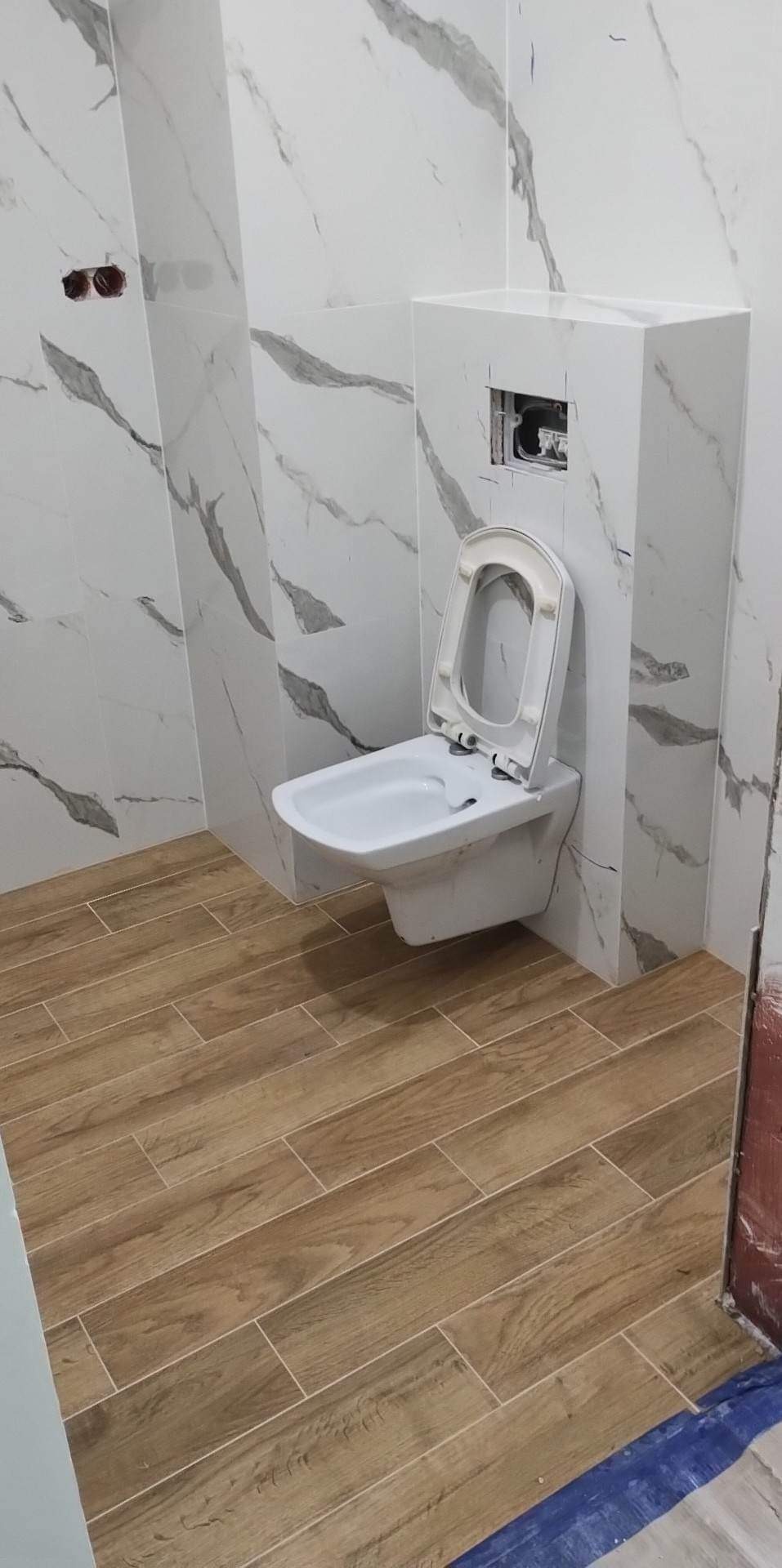Wisząca toaleta z otwartą klapą, obudowana płytkami imitującymi marmur, na podłodze płytki drewnopodobne, widoczne otwory na gniazdka elektryczne w ścianie.