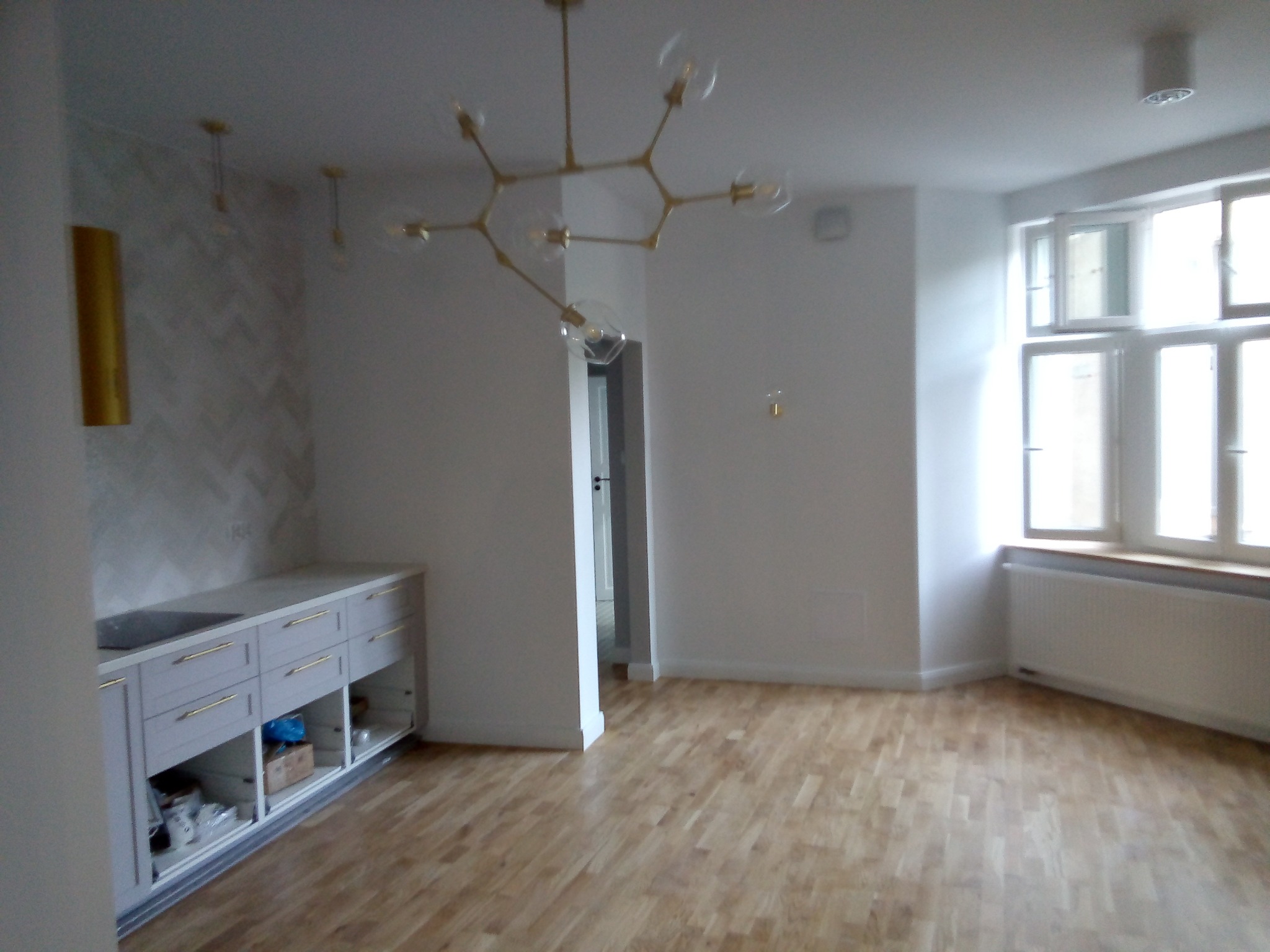 Jasne wnętrze po remoncie: parkiet, białe ściany, nowoczesna lampa sufitowa z kloszami, zabudowa kuchenna z szarymi szafkami i złotymi uchwytami, okno z białymi ramami, grzejnik.