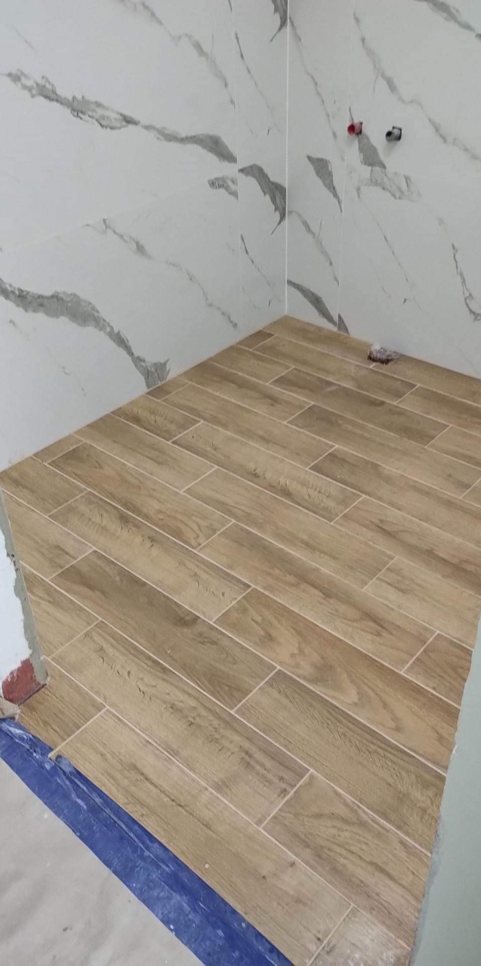 Ukończona podłoga w łazience wyłożona brązowymi płytkami imitującymi drewno, ściany wykończone płytkami marmurkowymi, widoczne fragmenty niezakończonych prac wykończeniowych.