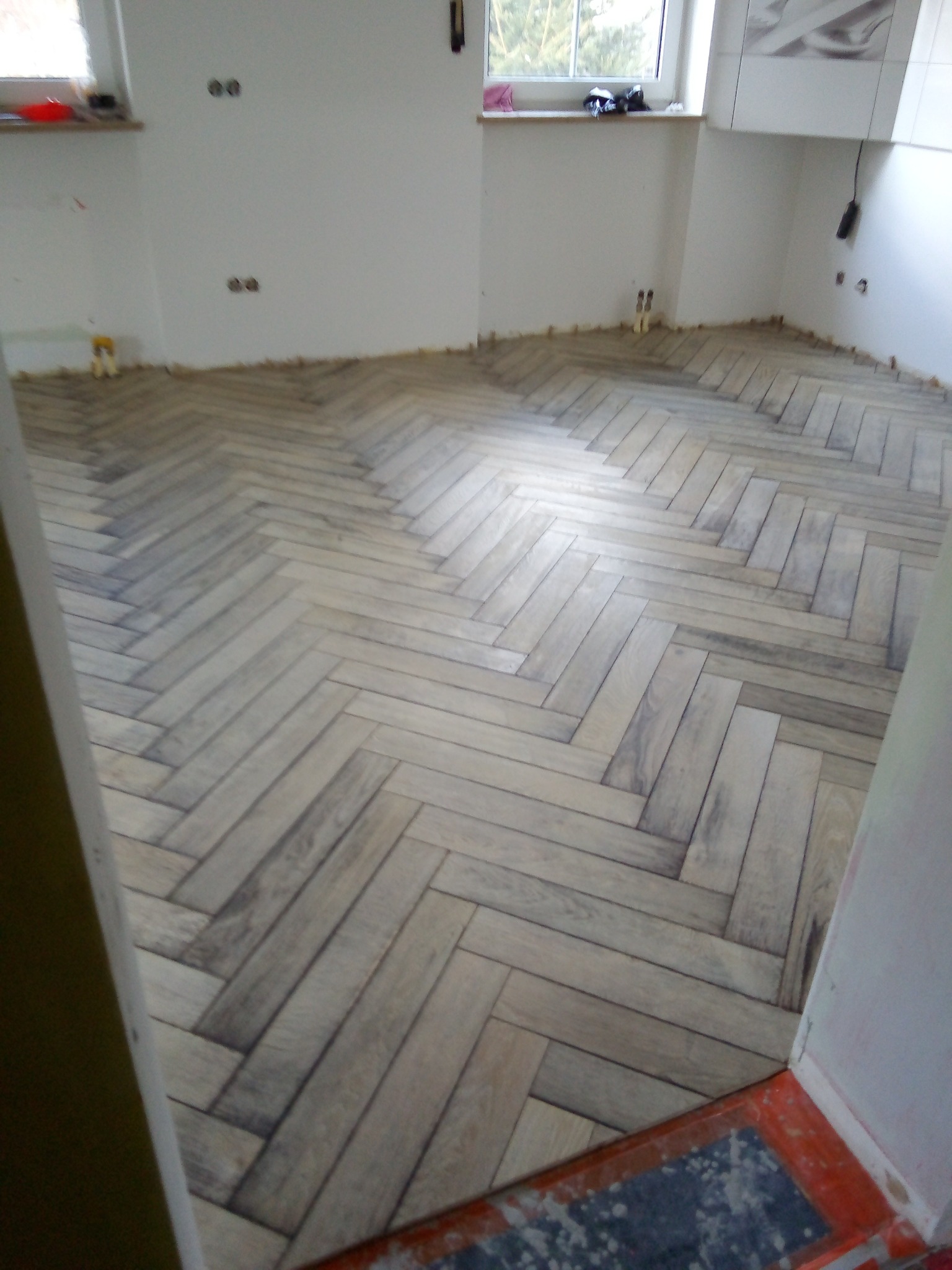 Ułożona w jodełkę podłoga z płytek imitujących drewno, widoczne niedokończone prace wykończeniowe przy ścianach.