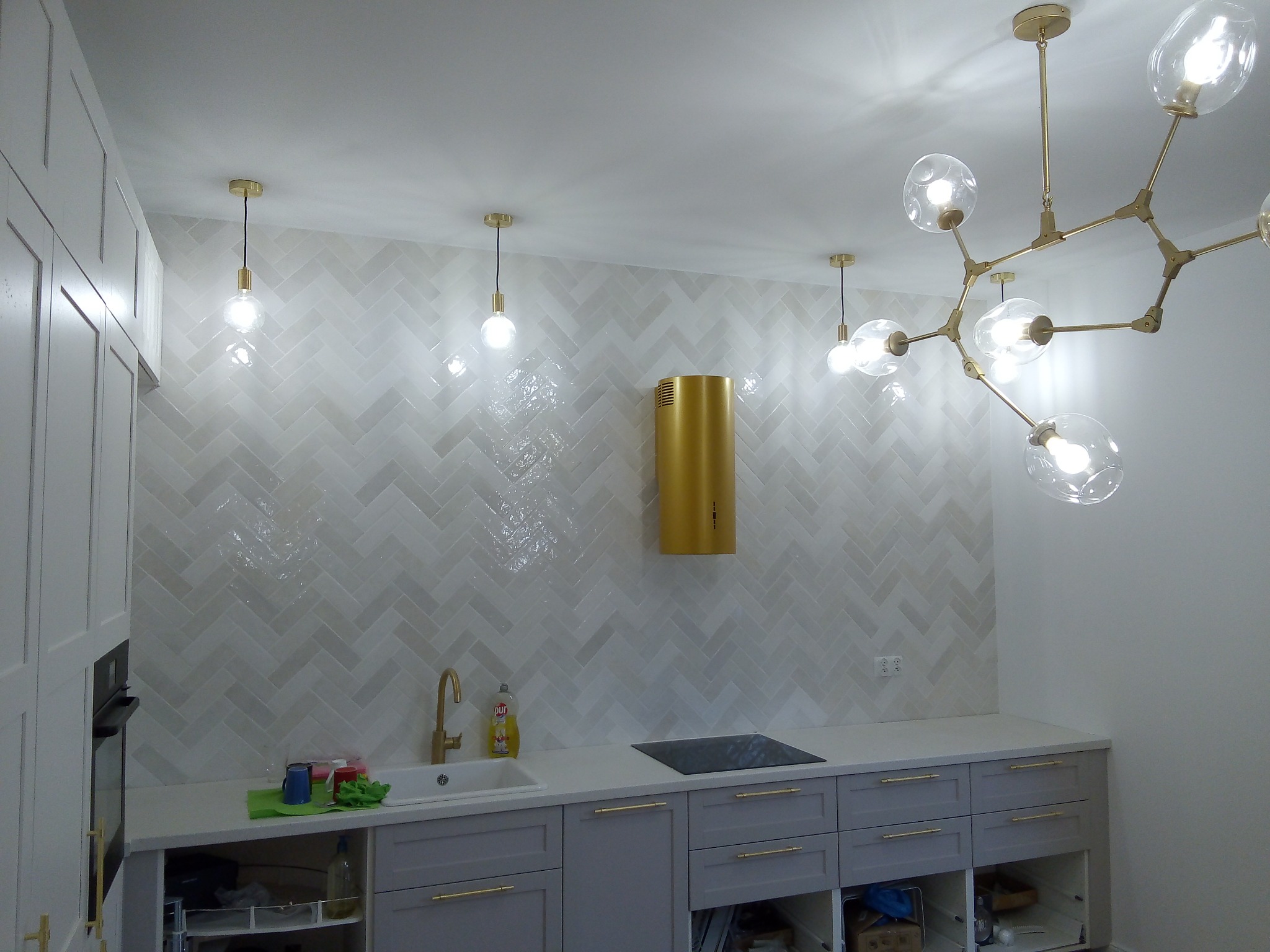 Nowoczesna kuchnia z szarymi szafkami, złotą armaturą i białymi płytkami ułożonymi w jodełkę na ścianie, złoty okap i designerska lampa sufitowa z żarówkami w kształcie kul.