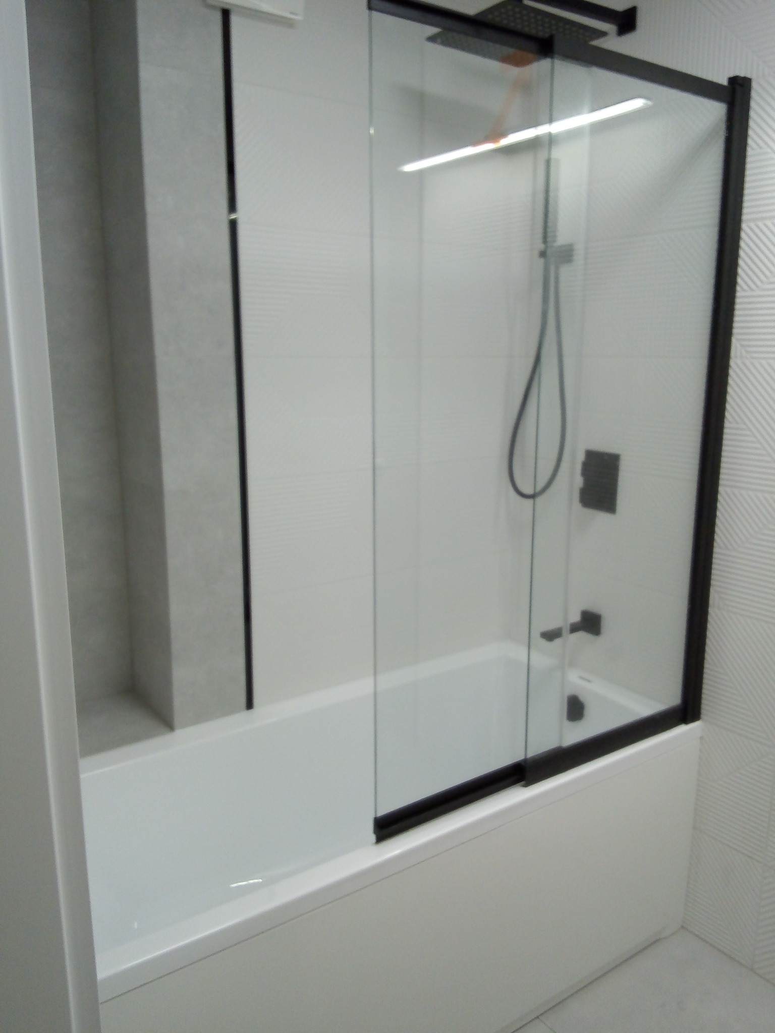 Nowoczesna łazienka z białą wanną obudowaną panelami, czarną szklaną kabiną prysznicową z przesuwnymi drzwiami i deszczownicą, oraz jasnymi płytkami na ścianach z delikatnym wzorem.