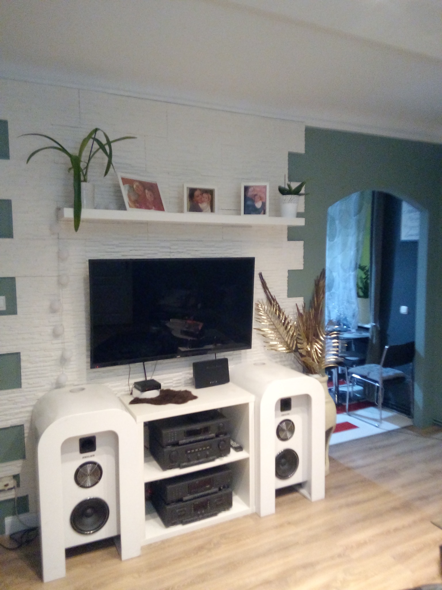 Pokój dzienny z telewizorem i białymi meblami na tle ściany w odcieniach bieli i szarości, z widocznym wejściem do innego pomieszczenia.