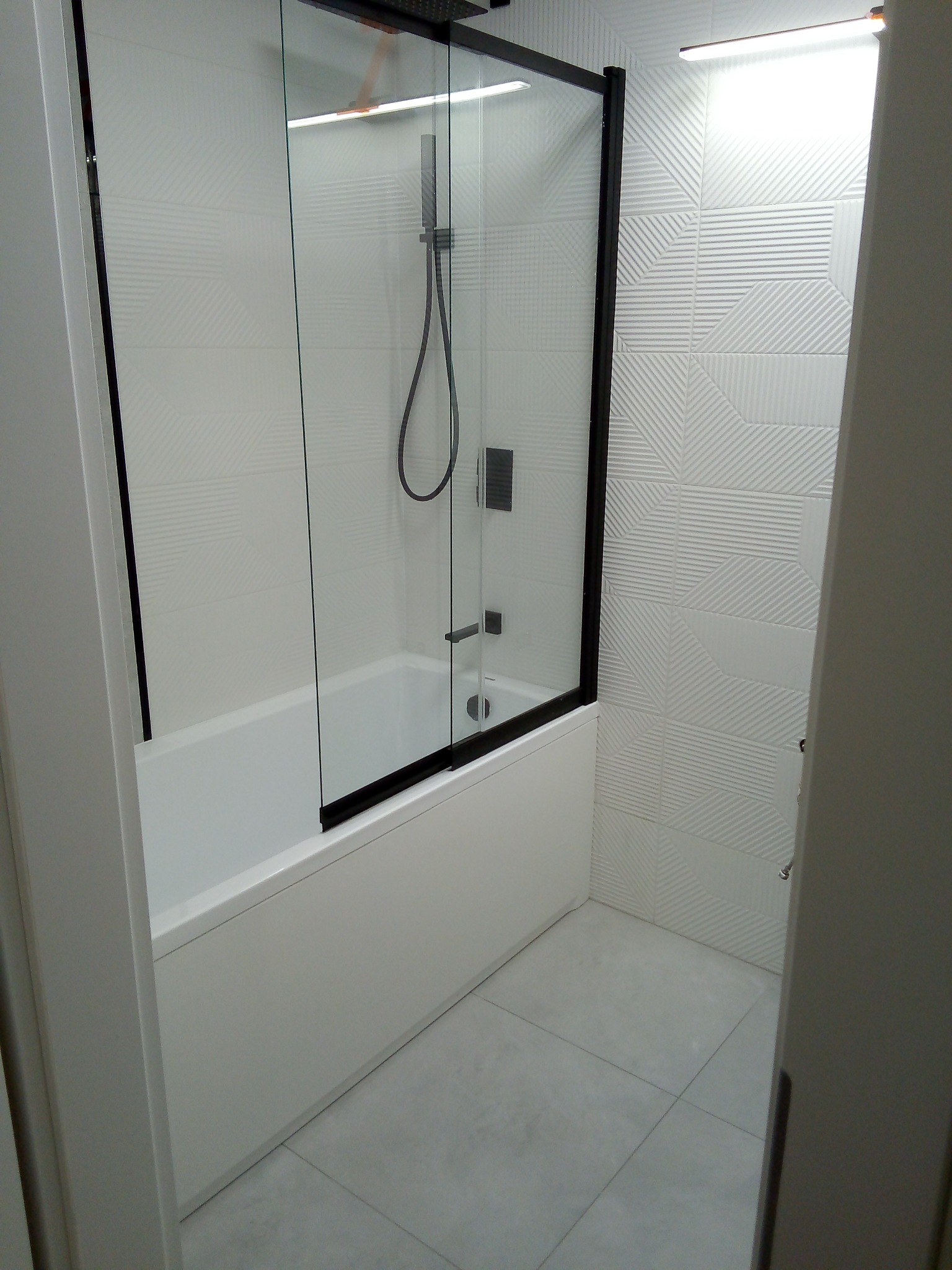 Nowoczesna łazienka z białą wanną, czarną ramą kabiny prysznicowej i geometrycznymi płytkami na ścianach.