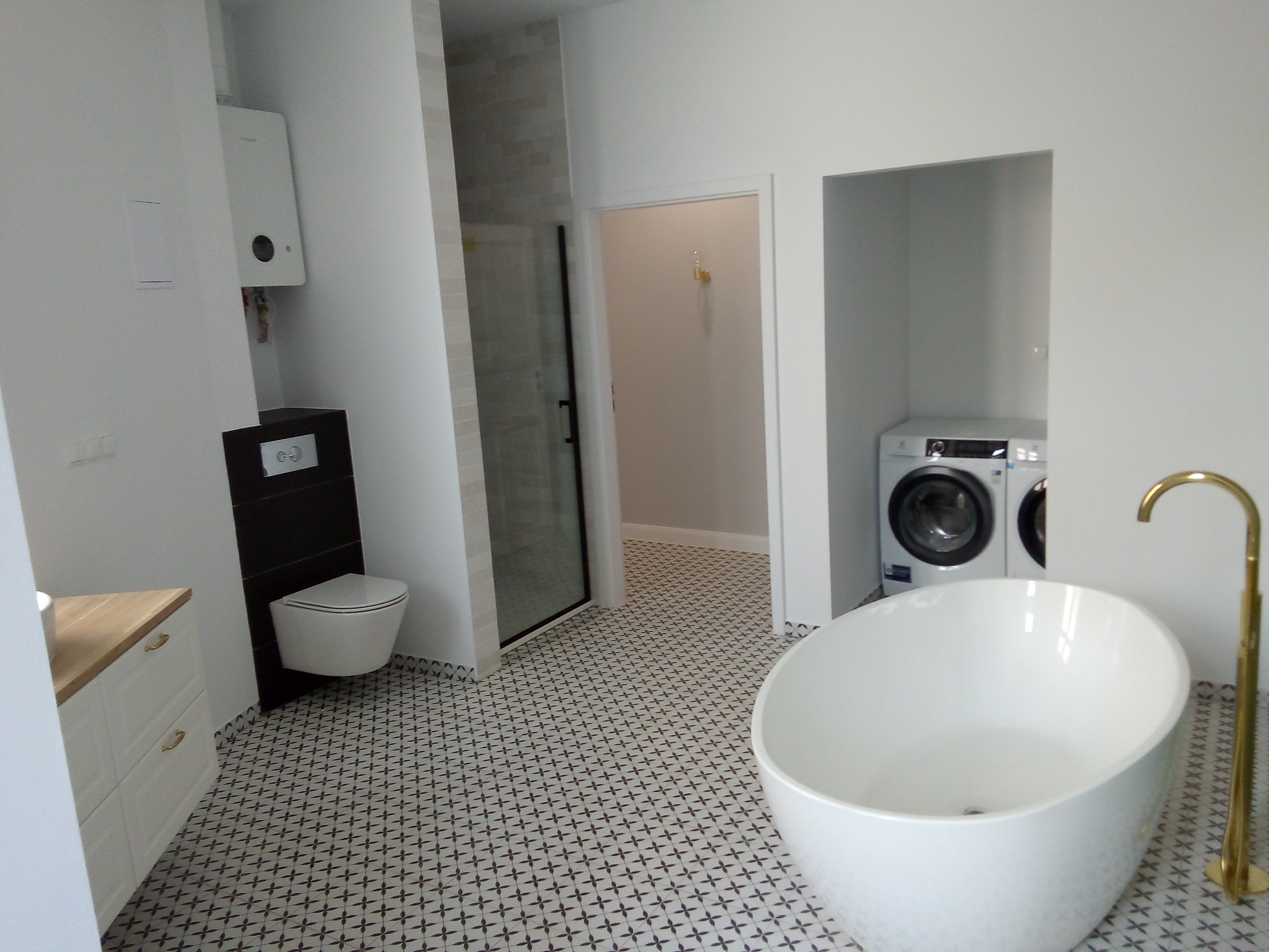 Nowoczesna łazienka z wolnostojącą wanną, złotą baterią podłogową, wiszącą toaletą, kabiną prysznicową typu walk-in i pralką zabudowaną we wnęce.