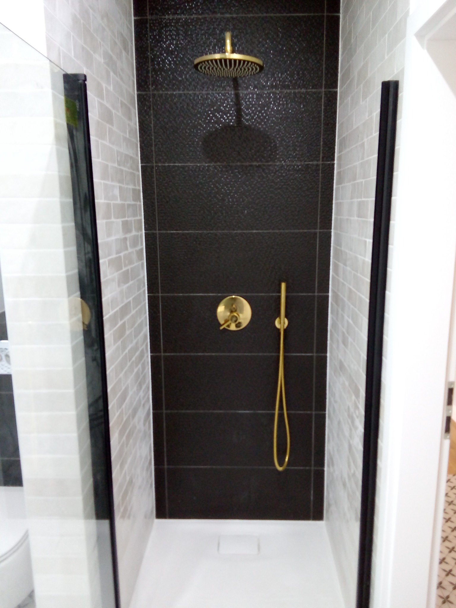 Nowoczesny prysznic walk-in z czarną ścianą z płytek o fakturze i złotą armaturą, otoczony jasnymi płytkami imitującymi cegłę i czarnymi ramami szklanych paneli.