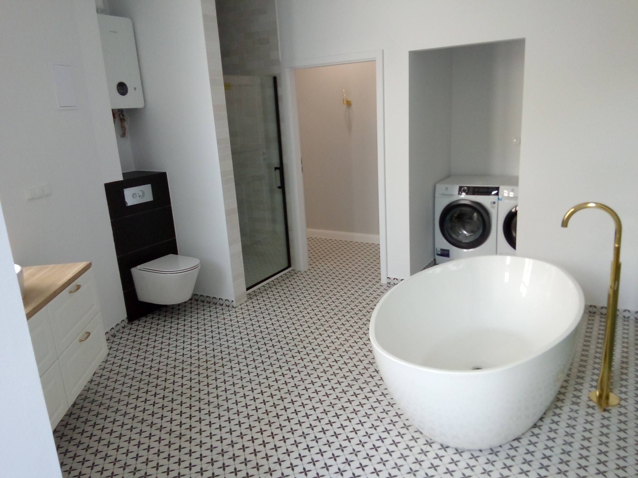 Nowoczesna łazienka z wolnostojącą białą wanną, złotą baterią, wzorzystą podłogą, toaletą podwieszaną i pralką w zabudowie.