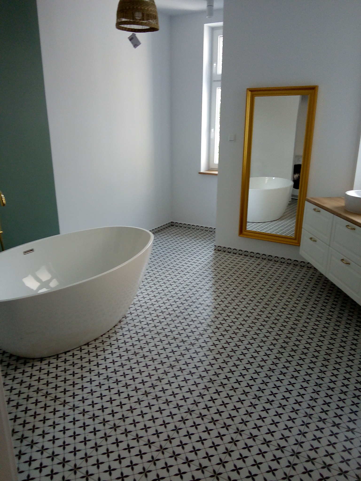 Nowoczesna łazienka z wolnostojącą białą wanną, wzorzystą podłogą w czarne krzyżyki, złotym lustrem i białymi szafkami z drewnianym blatem.
